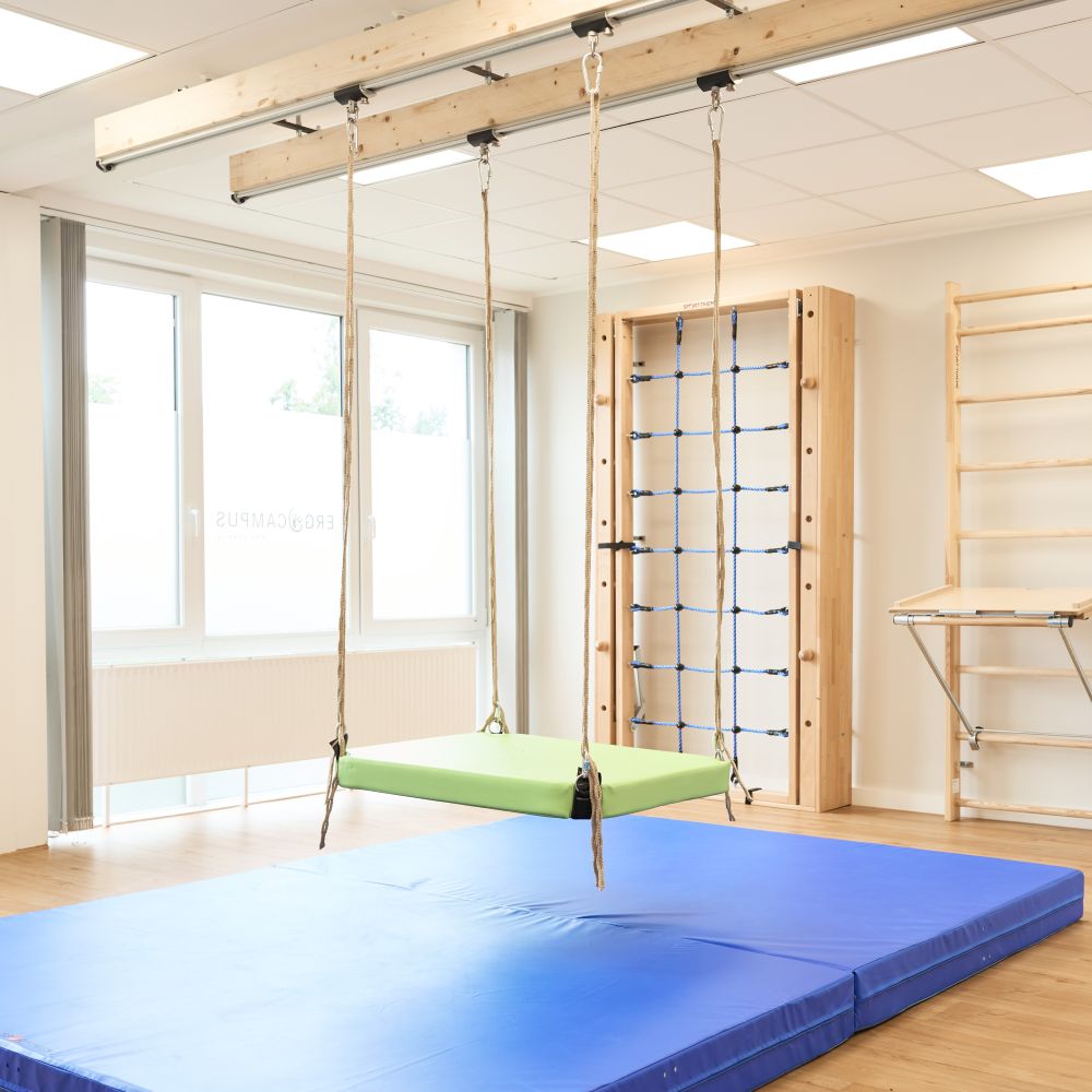 Therapieraum mit blauen Fallschutzmatten, einer an Seilen aufgehängten grünen Schaukel sowie Kletter- und Sprossenwandelementen aus Holz.